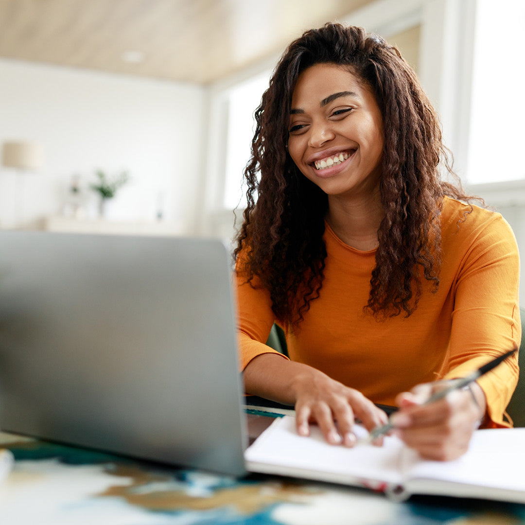 Woman sitting at a table with a laptop and notebook, smiling.