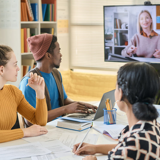 Three people in a meeting with a fourth person on a video call, in a room with books and a laptop.